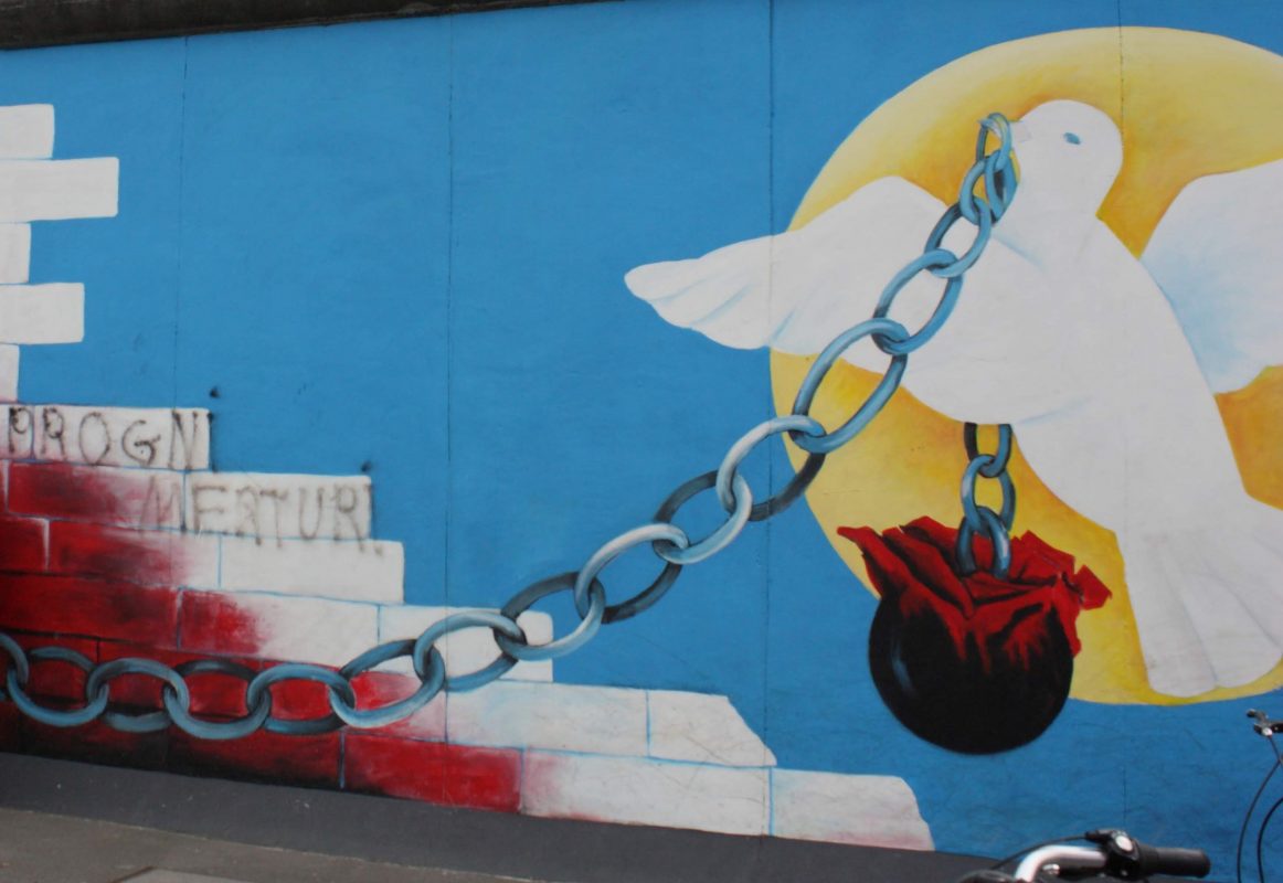 Kuva Berliinin muurista, jossa kyyhkynen pitää nokassaan kahletta, jonka päässä kukilla täytetty pata.