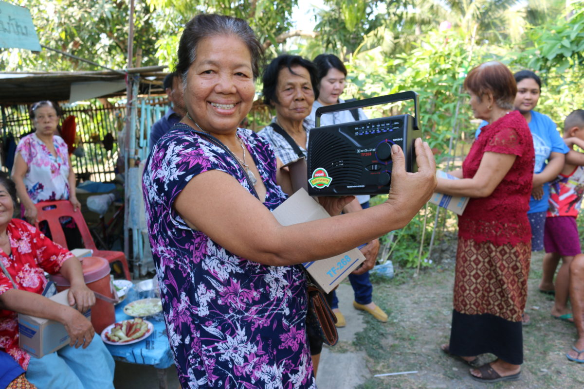 Iloisesti hymyilevä nainen näyttää kädessään olevaa radiota, jonka on juuri saanut lahjoituksena. Sen avulla on mahdollista kuunnella kristillistä opetusta.