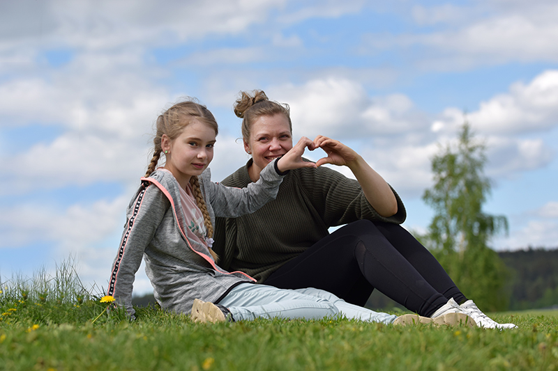 Kummitytär Lilli ja kummi Jenni istuvat nurmella ja tekevät sormillaan sydämen muodon.