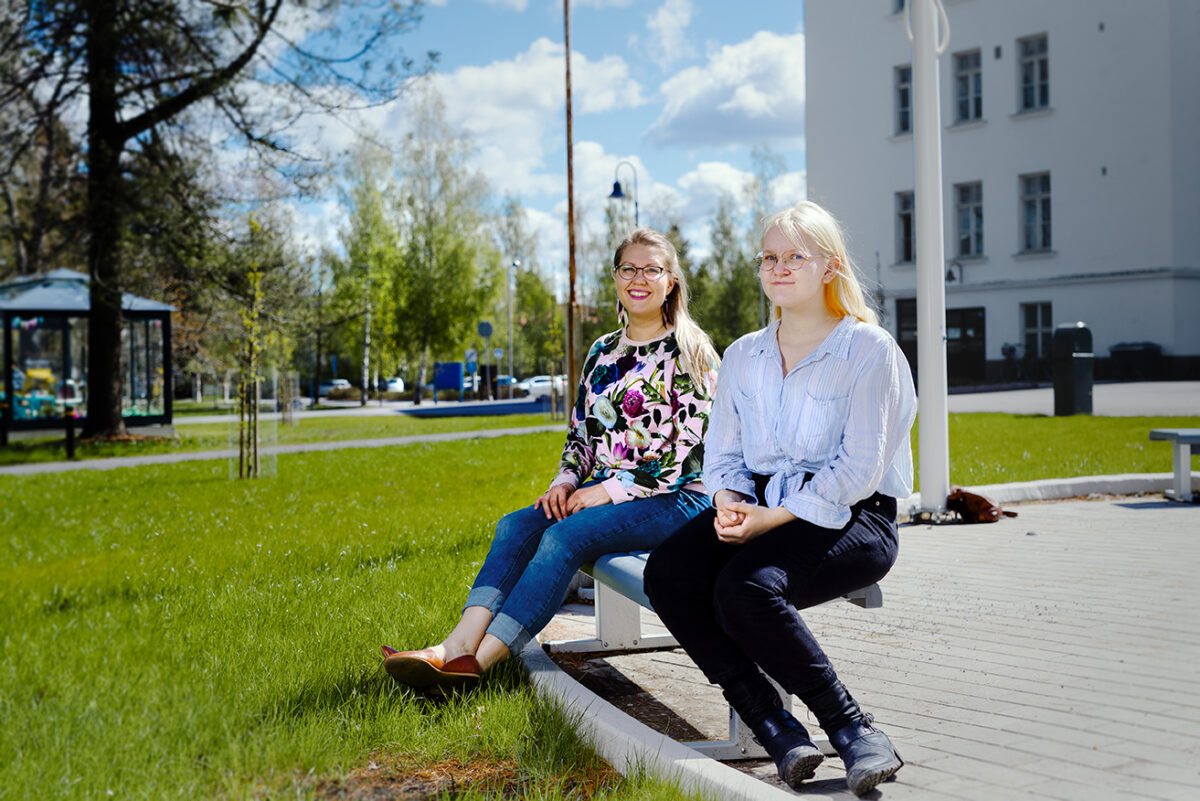 Kuvassa Anu Mäki-Latvala ja Miia Nortunen kesäisen nurmikon reunalla istumassa puiston penkillä.