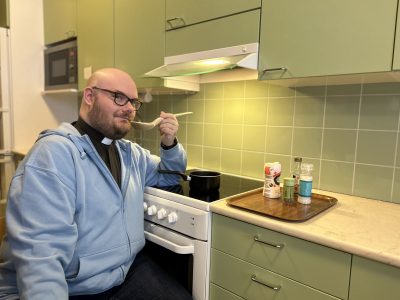 Vaaleansiniseen huppariin pukeutunut pastori istuu keittiössä lieden ääressä. Hellalla on pieni kattila, ja pastorilla on kädessään puukauha, josta hän on maistavinaan soppaa.