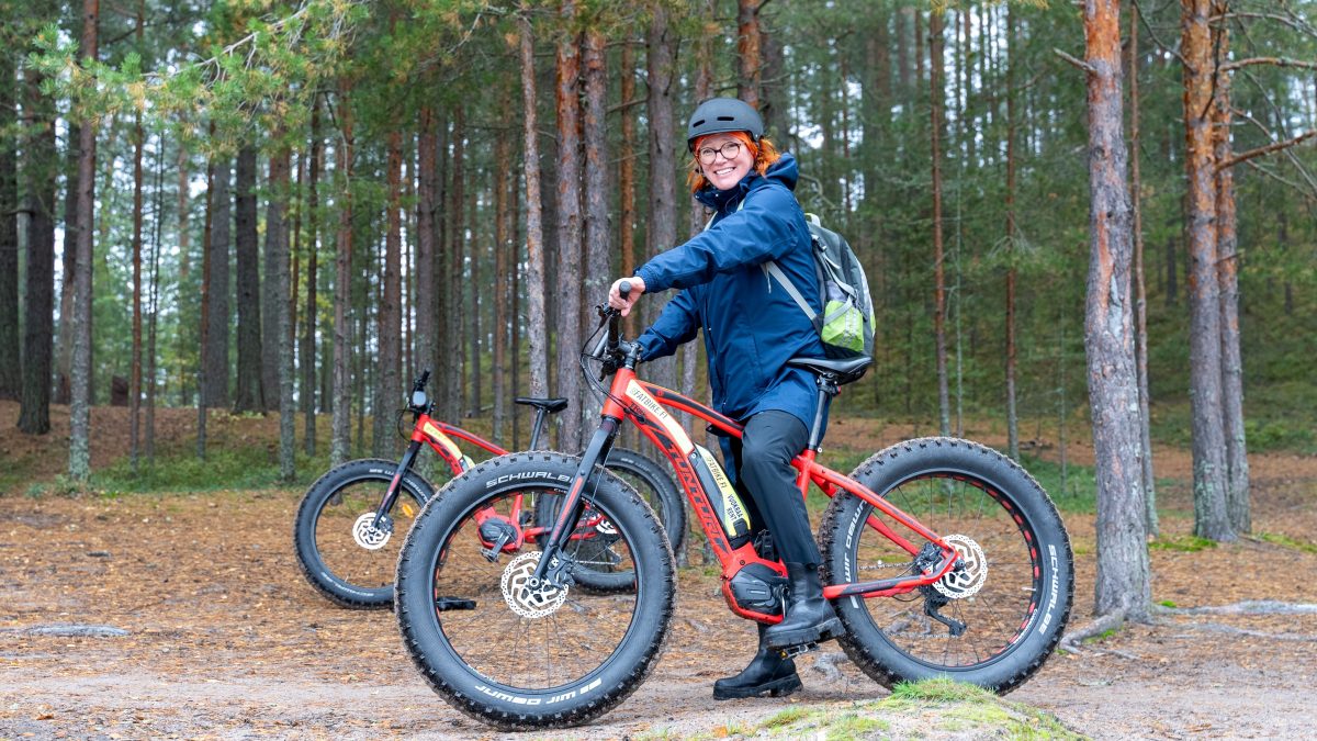 Piispa Mari Parkkinen pyörän selässä, takana mäntyistä kangasmetsää.