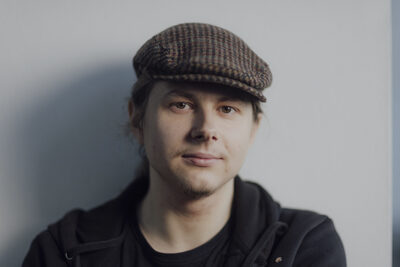 Olli Markonsaari katsoo vakavana kameraan. Päässä ruskea lippis, päällä musta huppari.