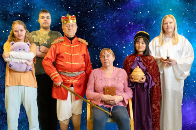 Ryhmä ihmisiä sinisen taivaan edessä. Yksi ihmisistä on pukeutunut kuninkaaksi ja yksi enkeliksi.