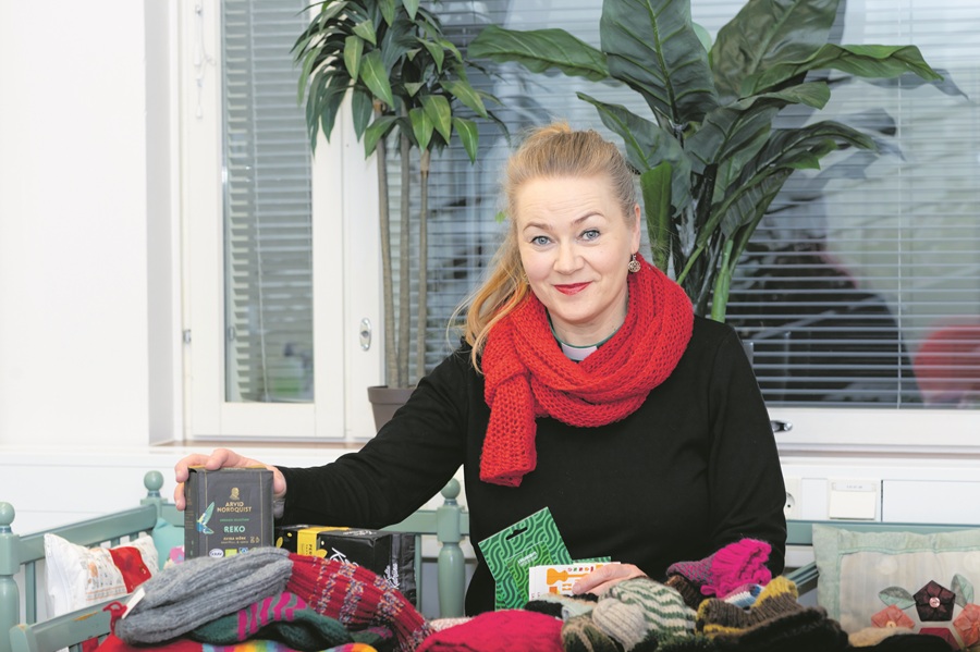 Punahuivinen nainen esittelee pöydän ääressä villasukkia, kahvipaketteja ja lahjakortteja.