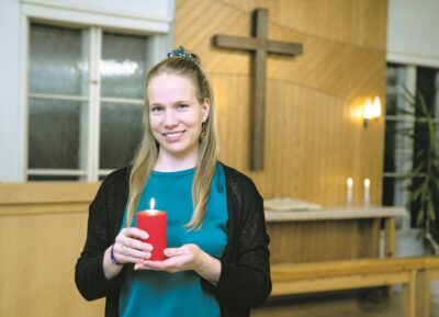 Vaaleahiuksinen nainen pitelee punaista kynttilää kirkossa.