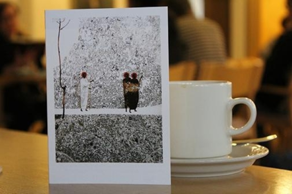 Kuvassa pöydällä oleva kahvikuppi ja sitä vasten nojaamassa postikortti, jossa ehkä afrikkalainen mukaelma Emmauksen tien kulkijoista.