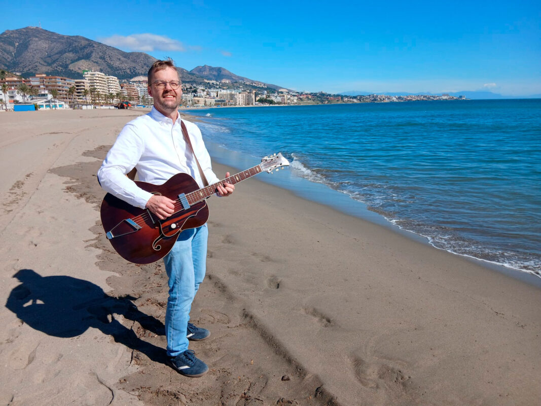 Kanttori Hannes Asikainen Espanjan Aurinkorannalla meren rannalla kitara käsissään.