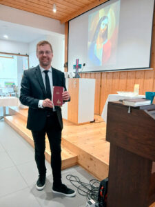 Hannes Asikainen kirkkotilassa virsikirja käsissään. Taustalla alttari ym. kirkon esineistöä. Kankaalle heijastettu piirroskuva Jeesuksesta. 