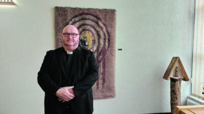 Silmälasipäinen miespappi seisomassa ryijyn edessä.