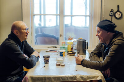 Timo Suutarinen istuu pöydän ääressä toisen miehen kanssa. Edessä on kahvikuppi ja pöydällä avonainen keksipaketti.