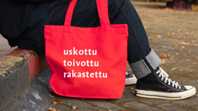 Ihminen istuu kadun reunakiveyksellä. Jalassa farkut ja tennarit. Vieressä punainen kassi, jossa valkoisella teksti: uskottu, toivottu, rakastettu.